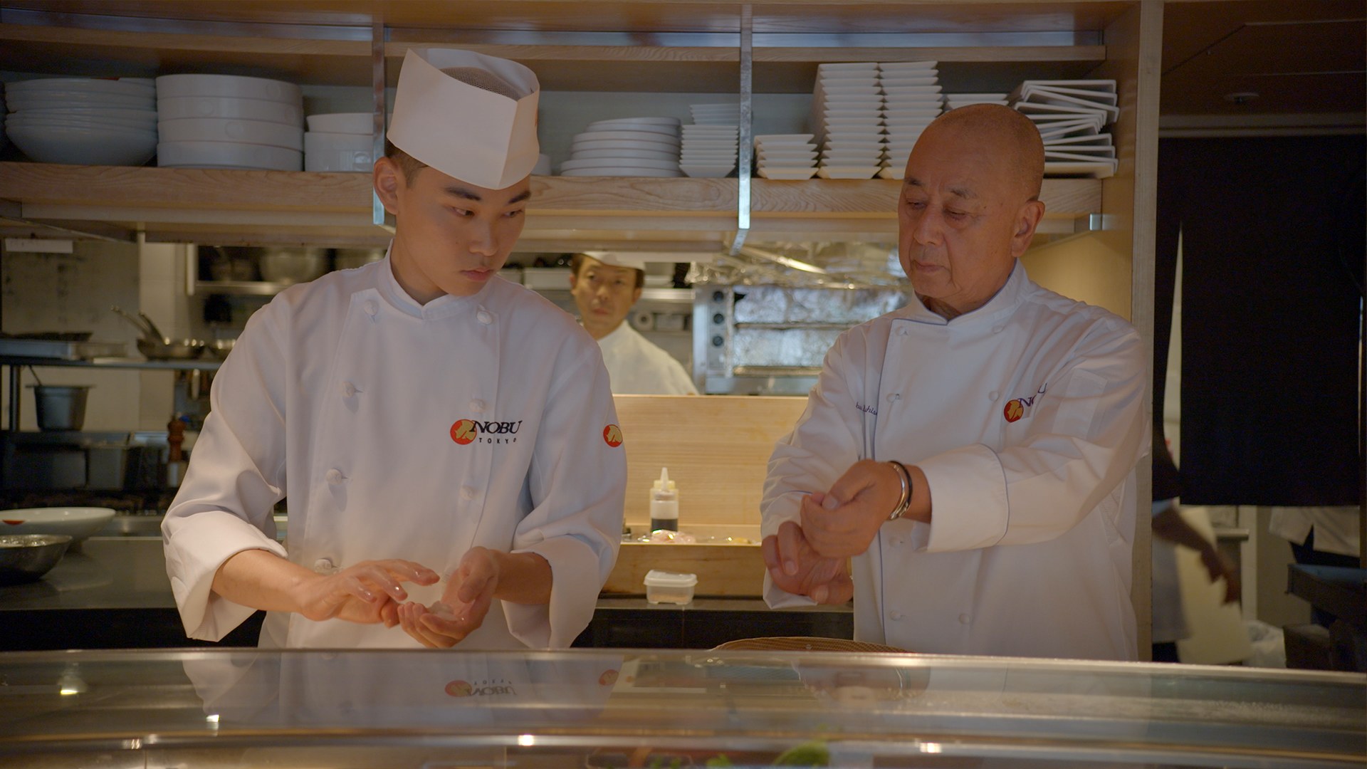 A senior chef works beside a young sushi chef at a restaurant counter in a still from the documentary Nobu.