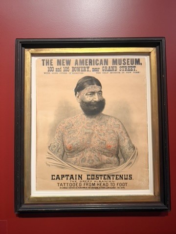 Framed nineteenth-century poster on a dark red wall reading The New American Museum, 103 and 105 Bowery, near Grand Street, with a portrait of a tattooed, bearded man billed as Captain Costentenus