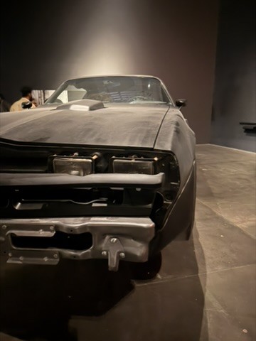 A full-size black car with its front grille stripped back, parked on a polished concrete floor in a dark gallery