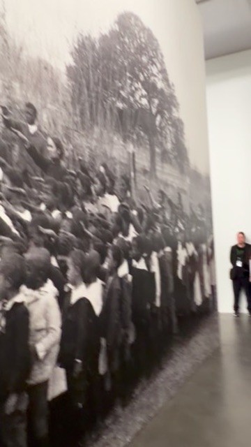 A floor-to-ceiling black-and-white photomural of a large crowd beneath trees, with a visitor standing at its edge for scale