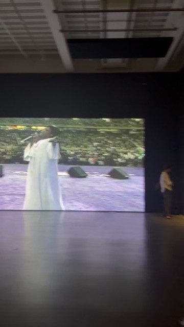 A large video projection in a dark gallery showing a figure in a long white garment standing outdoors, with a visitor silhouetted at right